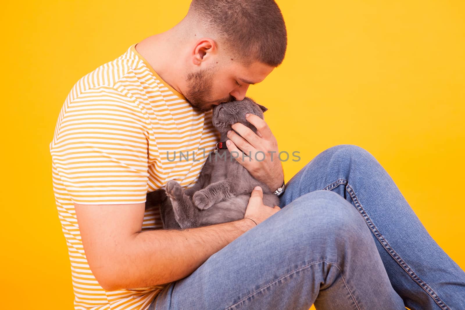 Handsome young man kissing and showing affection to his cat in studio over yellow background. Man loving his cat.