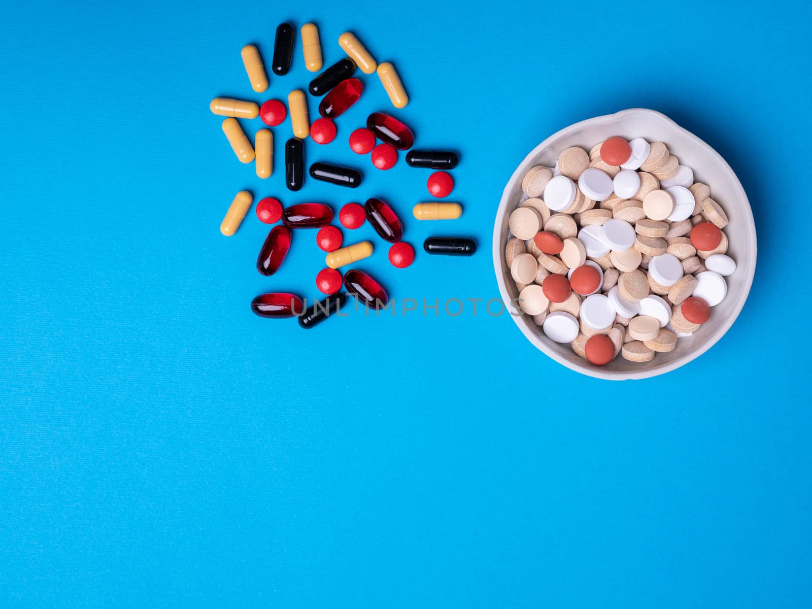 Top view with a bowl of pills and next to other pills and capsules of different colors and shapes. Copy space and blue background