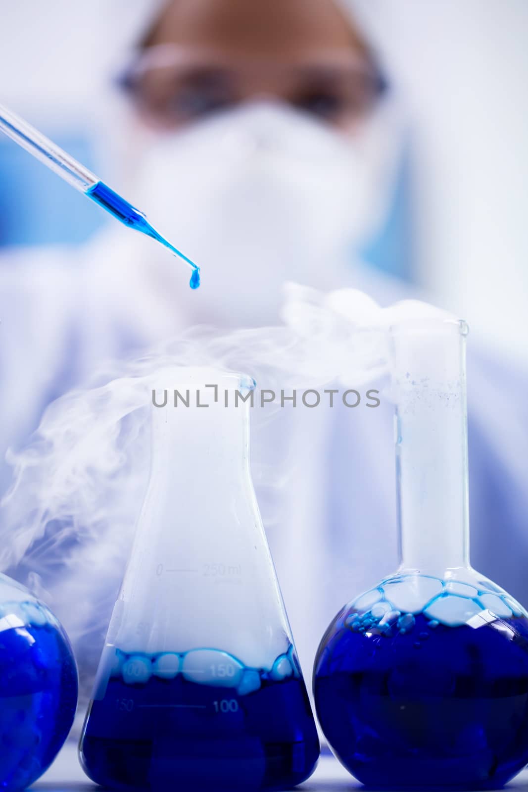 Close up of blue solution dropping from pipette in a test tube with smoking liquid. Scientist in protection equipment.