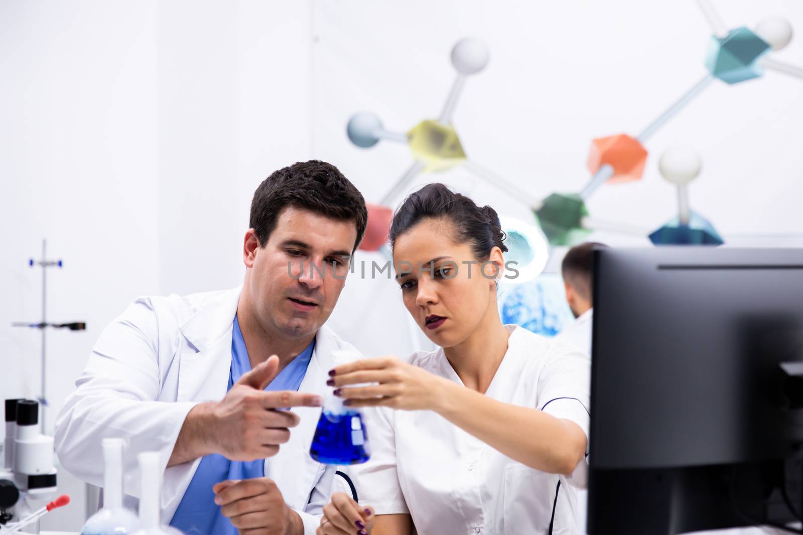 Scientist in white coat and his assitant looking at the bottle with blue liquid in modern research laboratory.