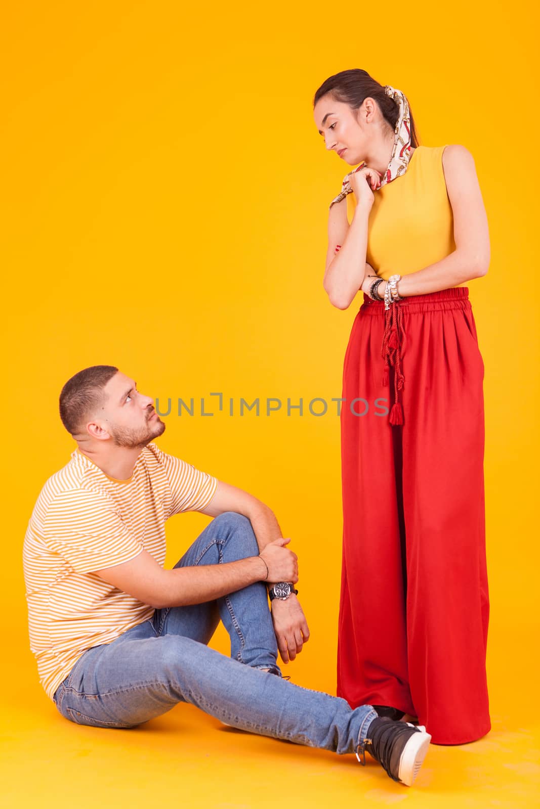 Confident boyfriend looking at his beautiful girlfriend sitting down over yellow background.