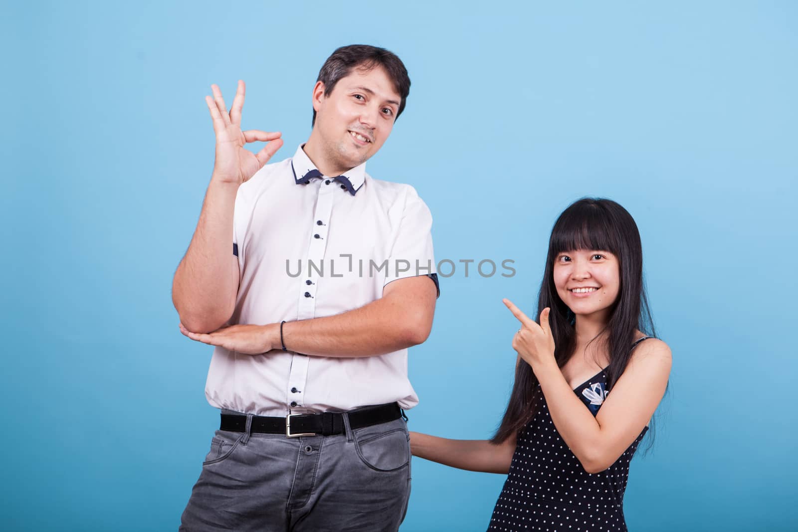Smiling asian girlfriend pointing at his caucasian boyfriend while he shows ok hand gesture. Interracial couple.