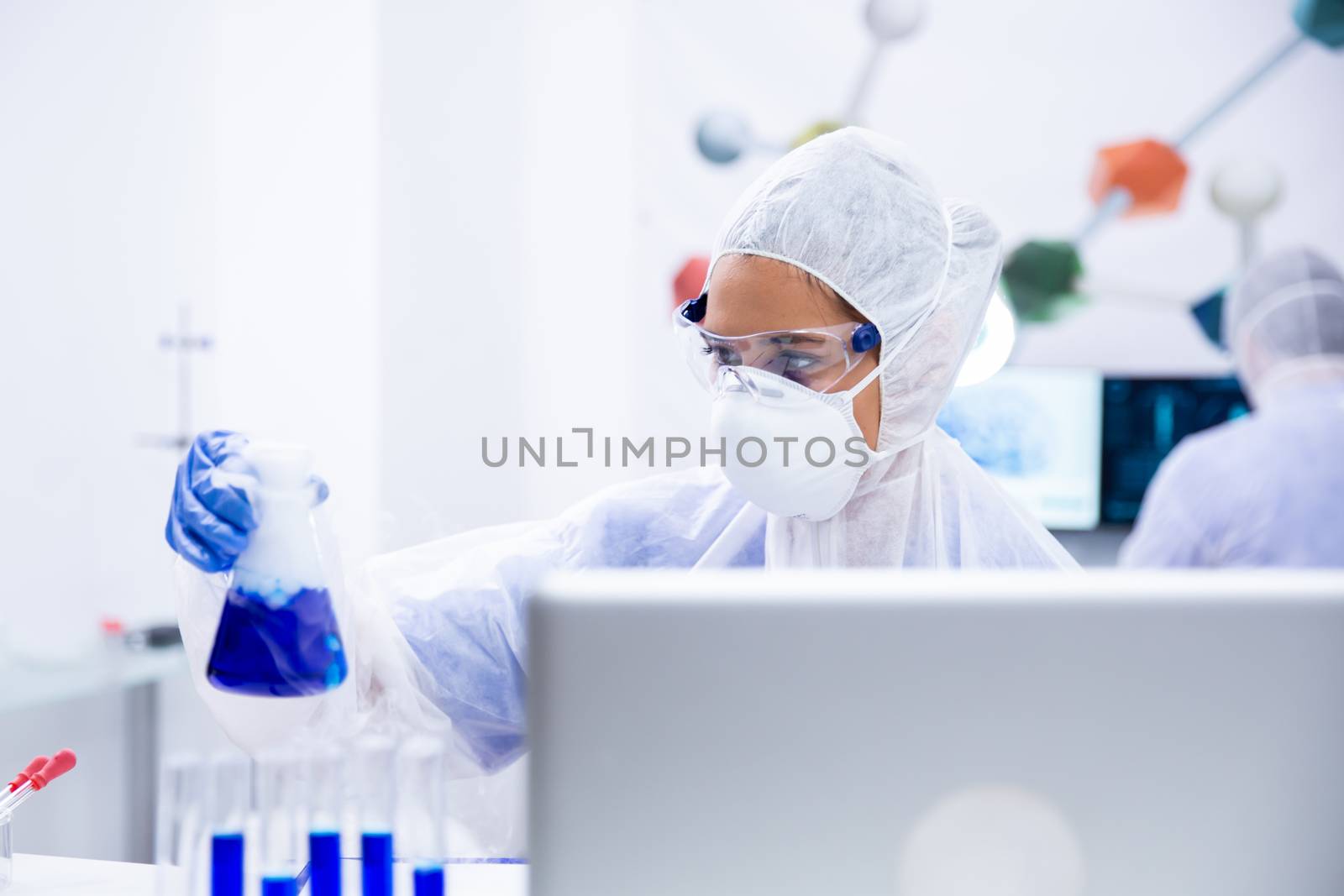 Researcher looks at a blue liquid container in front of a laptop. Glasses and protective suit
