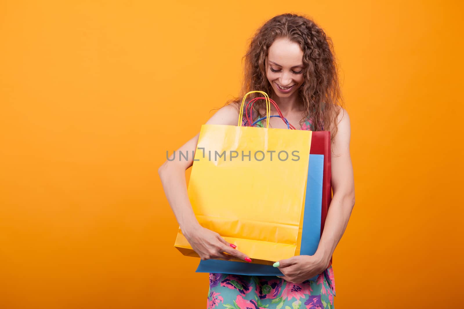 Gorgeous woman smiling and looking down on her colorful shopping bags over orange background in studio.