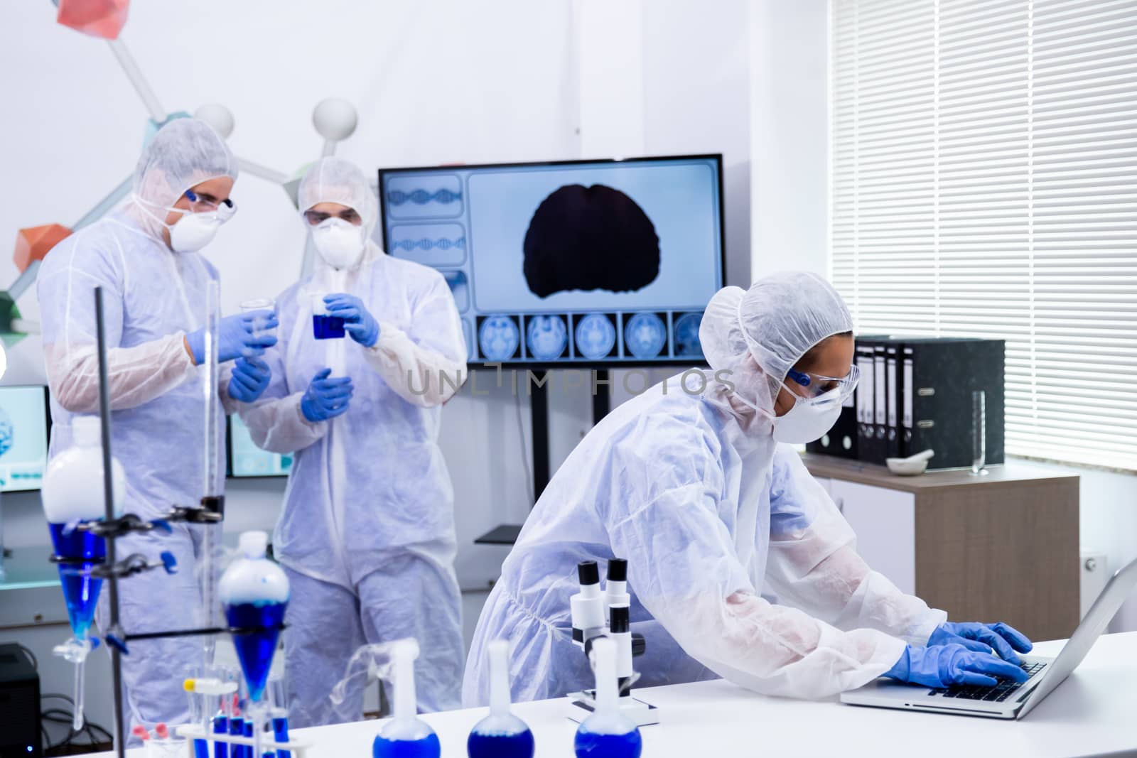Female scientist working on laptop and colleagues working with reactive blue solution in a modern research laboratory.