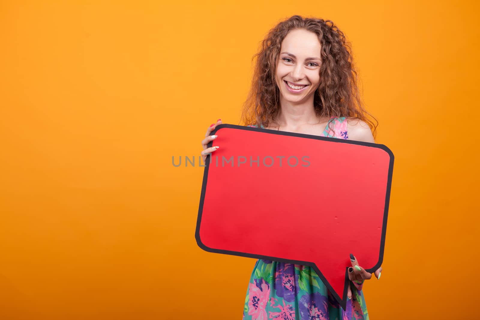 Friendly woman smiling and holding a blank sign in studio over orange background.