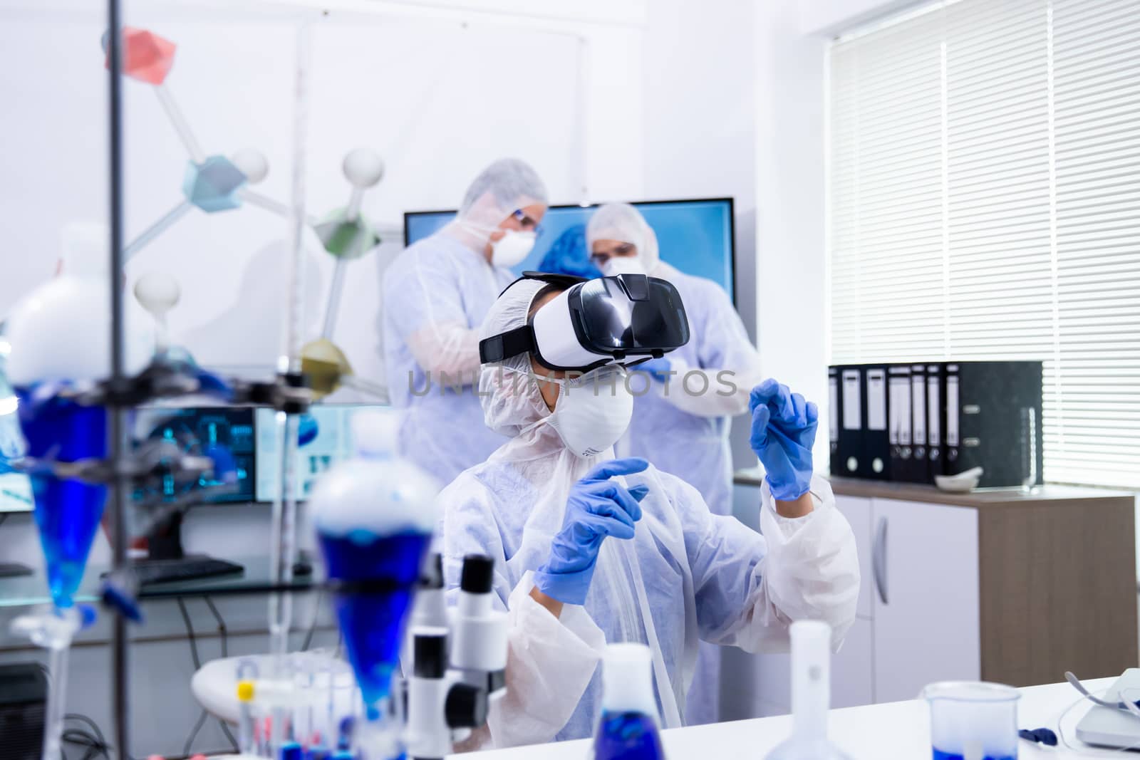 Scientist with virtual reality glasses doing test and her assistants working in the background with reactive blue liquid.