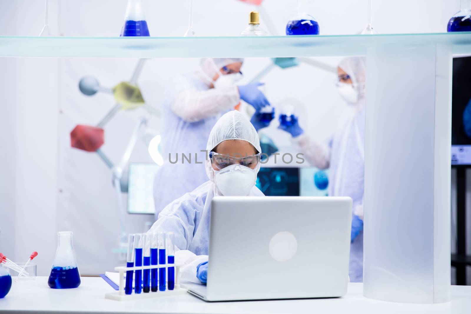 Chemist stands in front of the laptop with some test tubes next to him and two colleagues discuss in the back