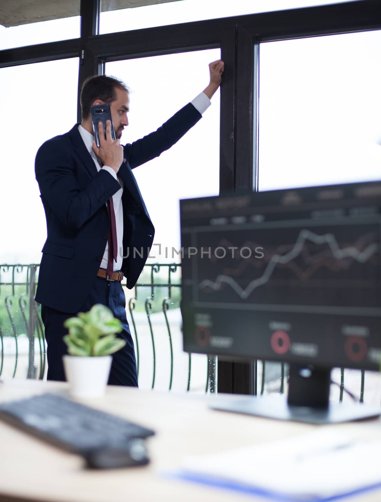 Handsome businessman in suit talking on the phone and looking on the window from his office. Businessman standing.