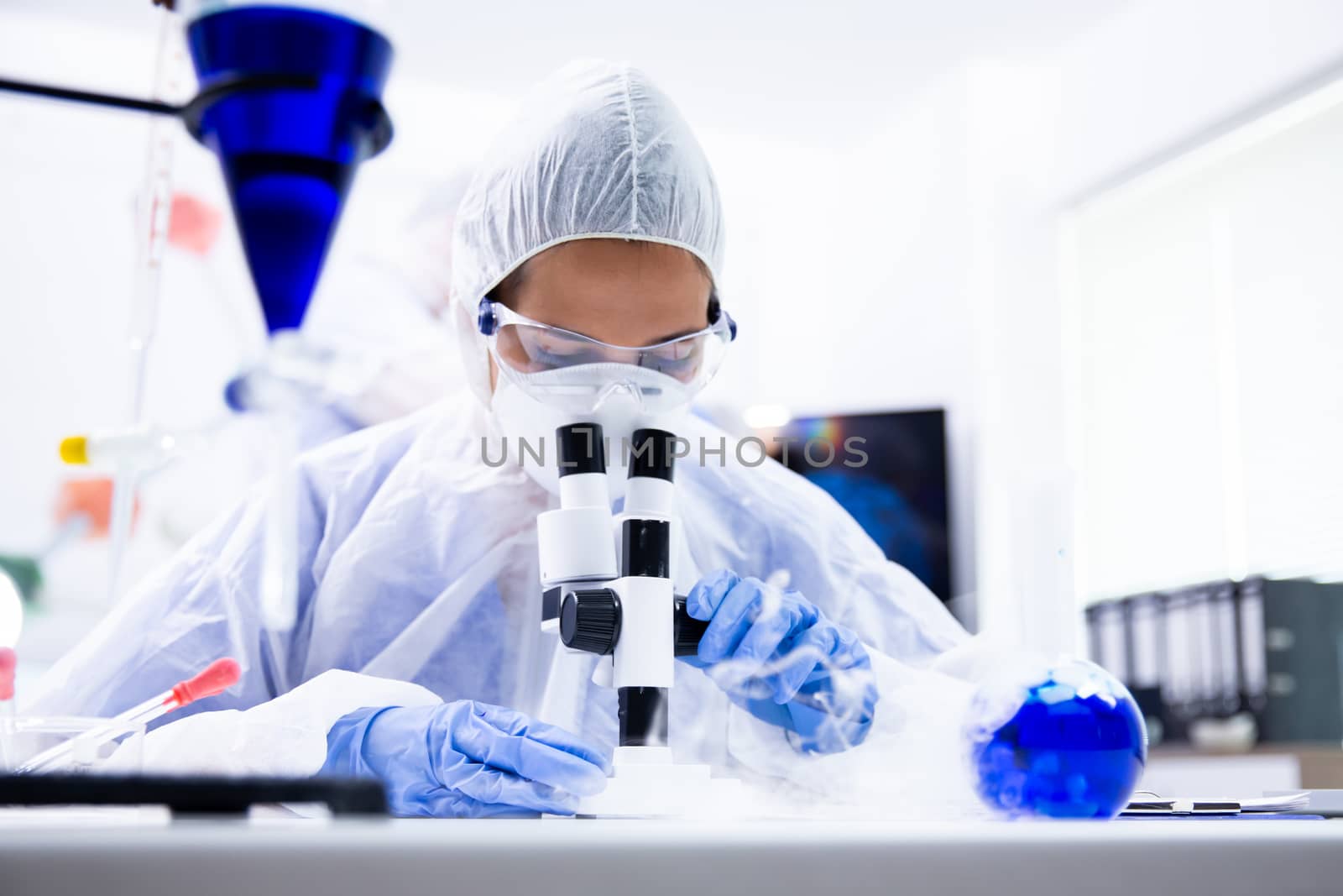 The researcher looks under the microscope near a beaker from which white smoke comes out. Blue glove and goggles
