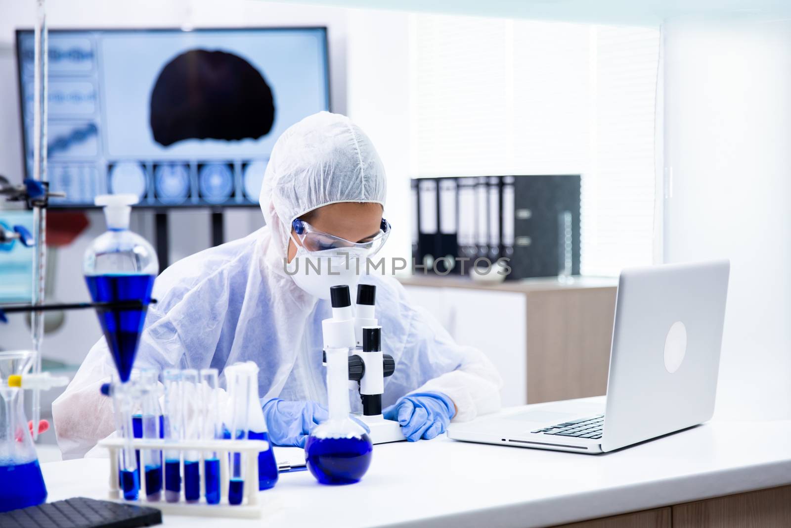 Caucasian chemist who looks under the microscope and is surrounded by many tubes. Blue substance and laptop