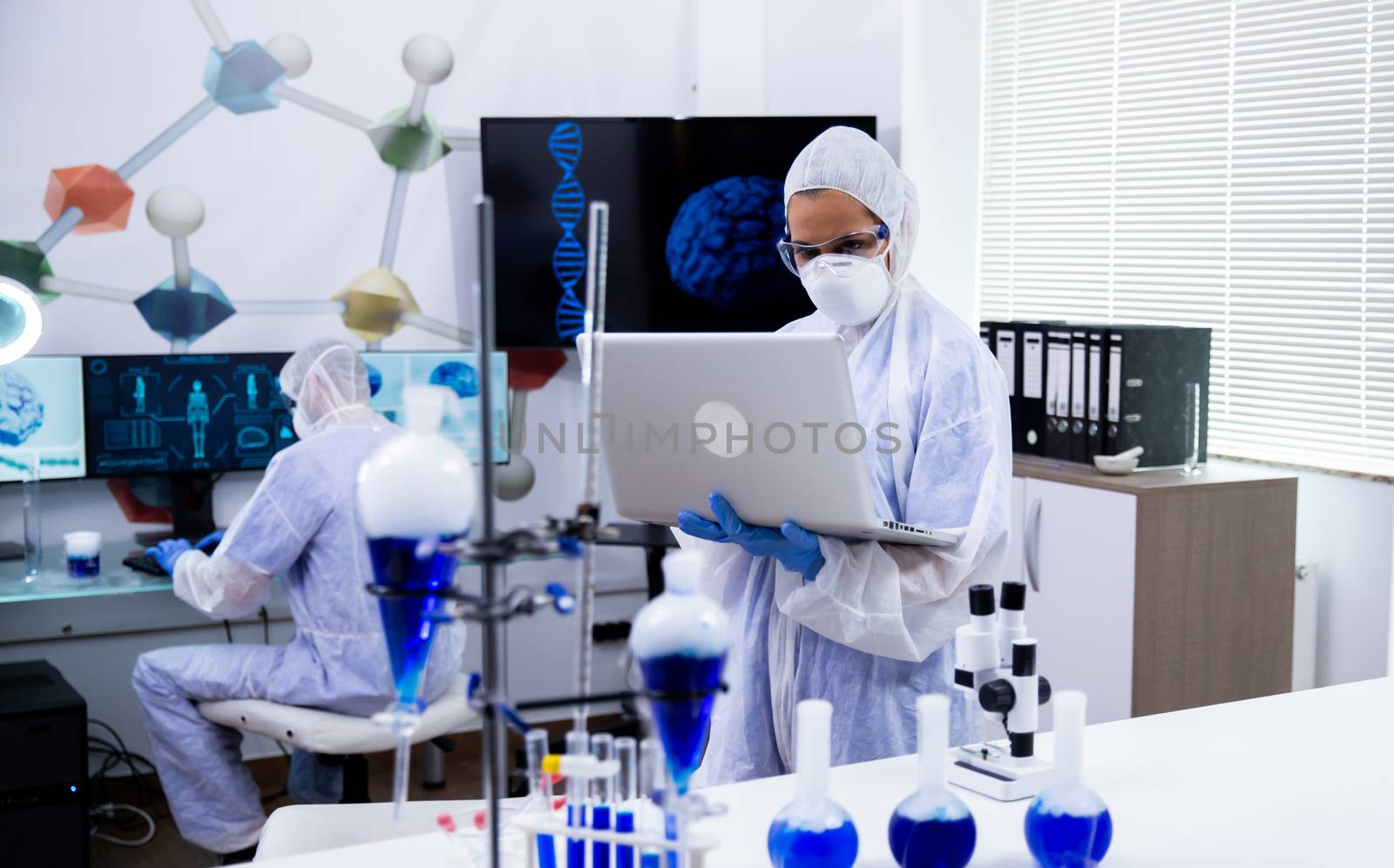 Scientist in modern research laboratory standing and working on laptop wearing protection for smoking blue soltion in test tubes.