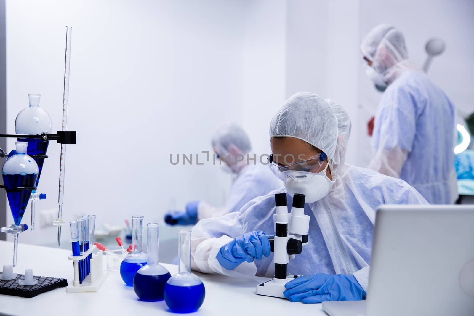 Female scientist looking at a sample of solution through microscope and team of researchers in the background.