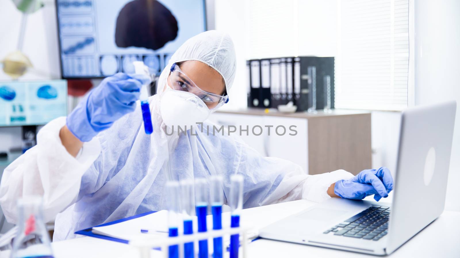 Caucasian scientist analyzes the tube and make some notes on laptop. Blue gloves and goggles