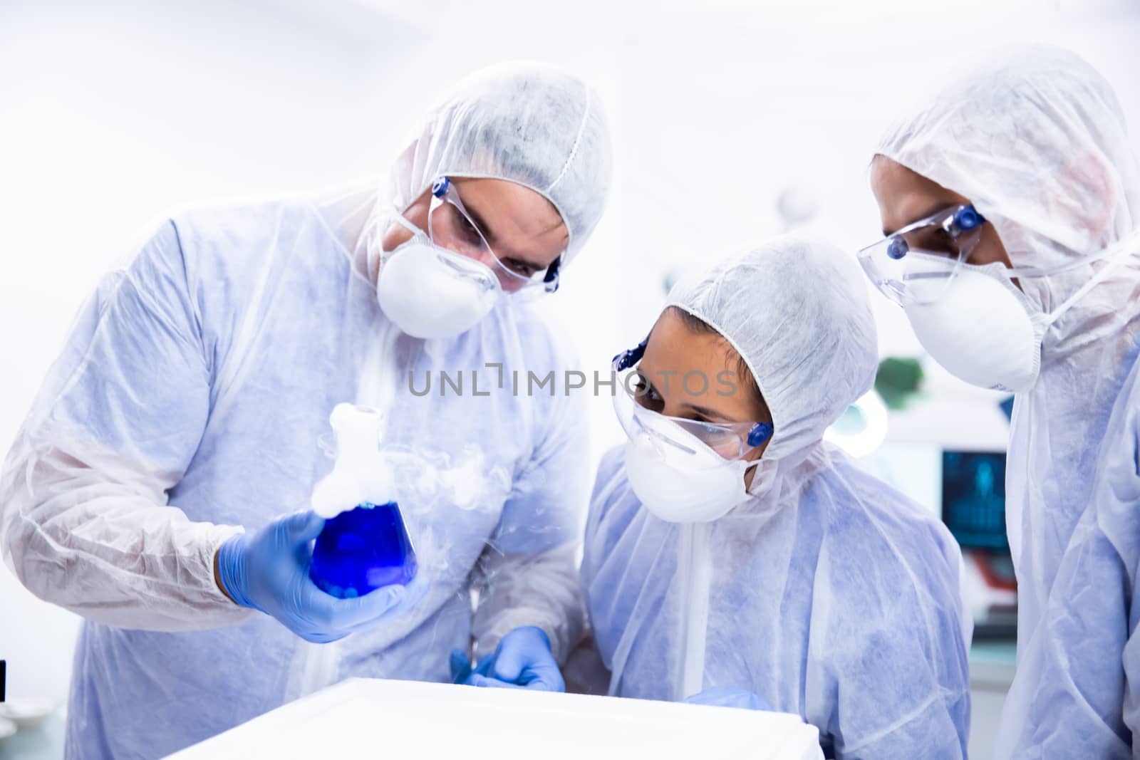 Scientist holds a beaker in his hand and two colleagues follow the reaction. Blue substance and white smoke