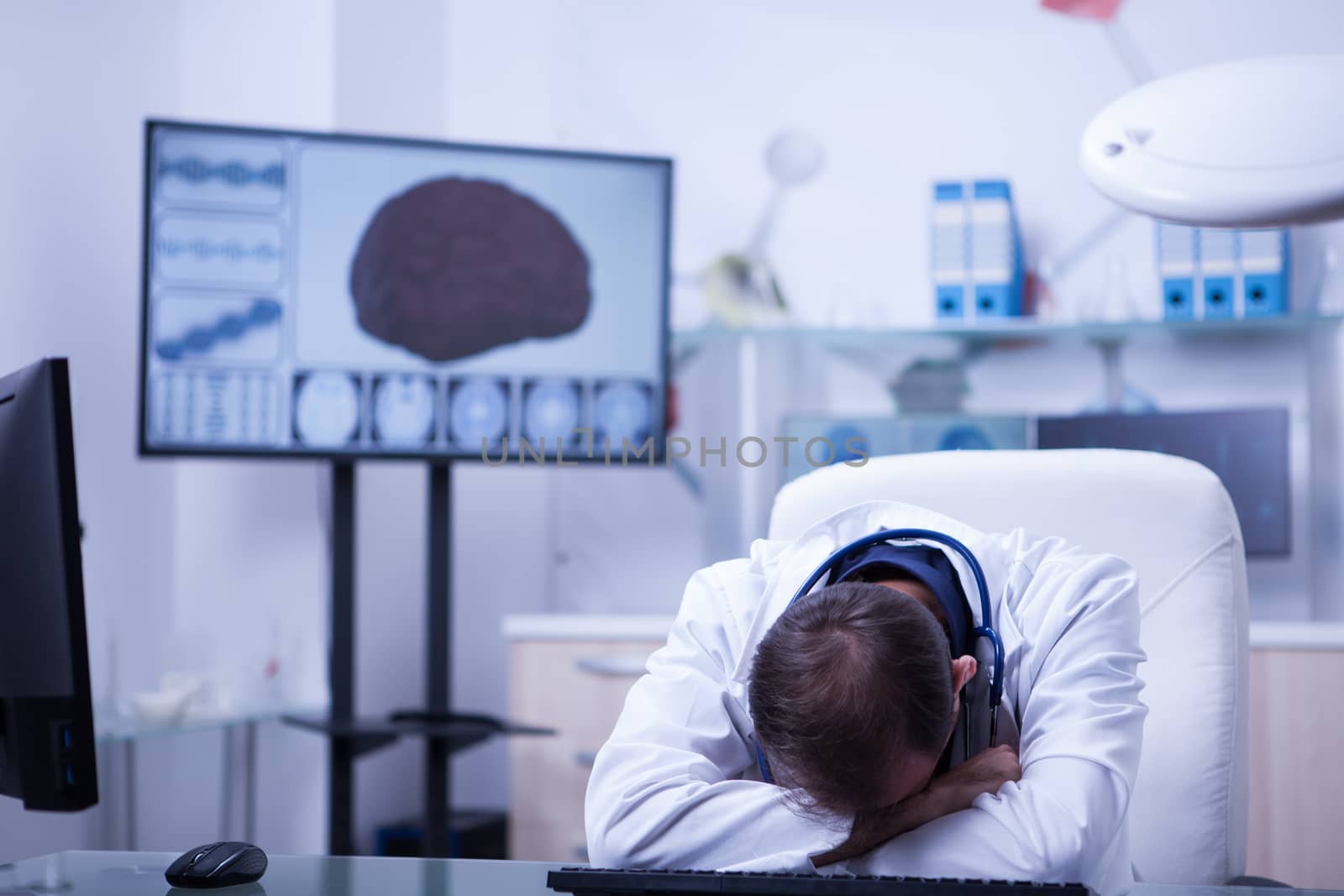 Young caucasian doctor sitting at this desk in the hospital with his heand on the desk sleeping. Overwokerd doctor.