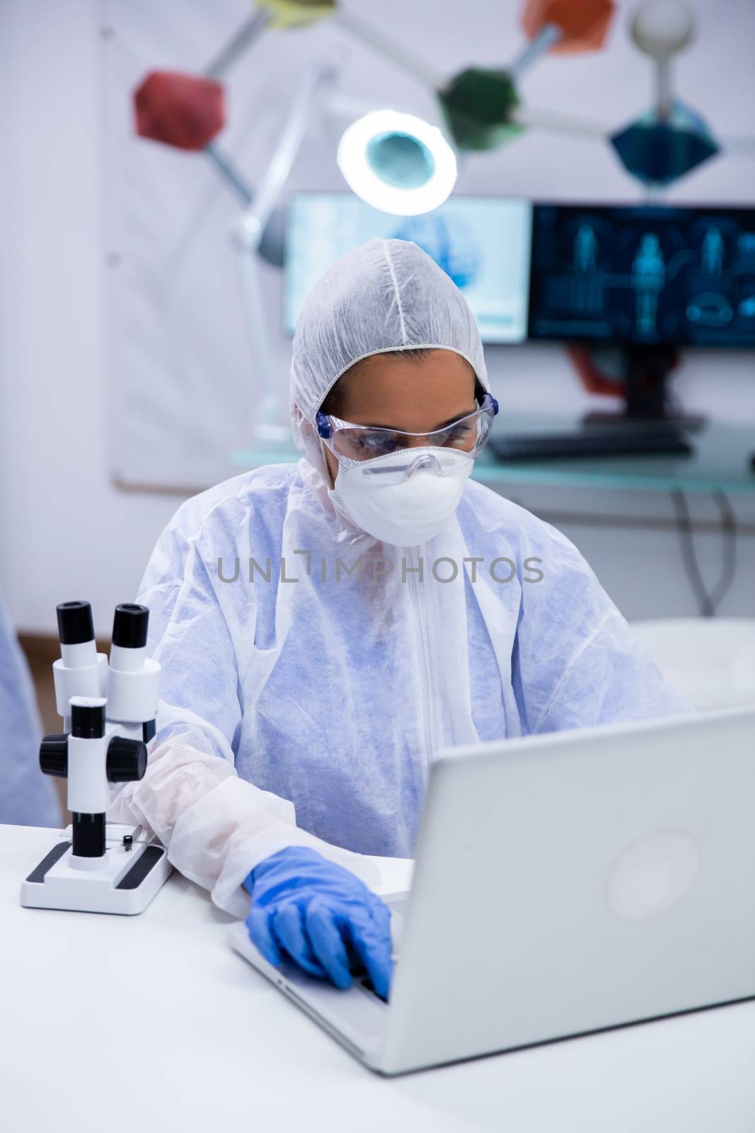 Focused female scientist wearing protection equipment working on her laptop in a modern research laboratory.