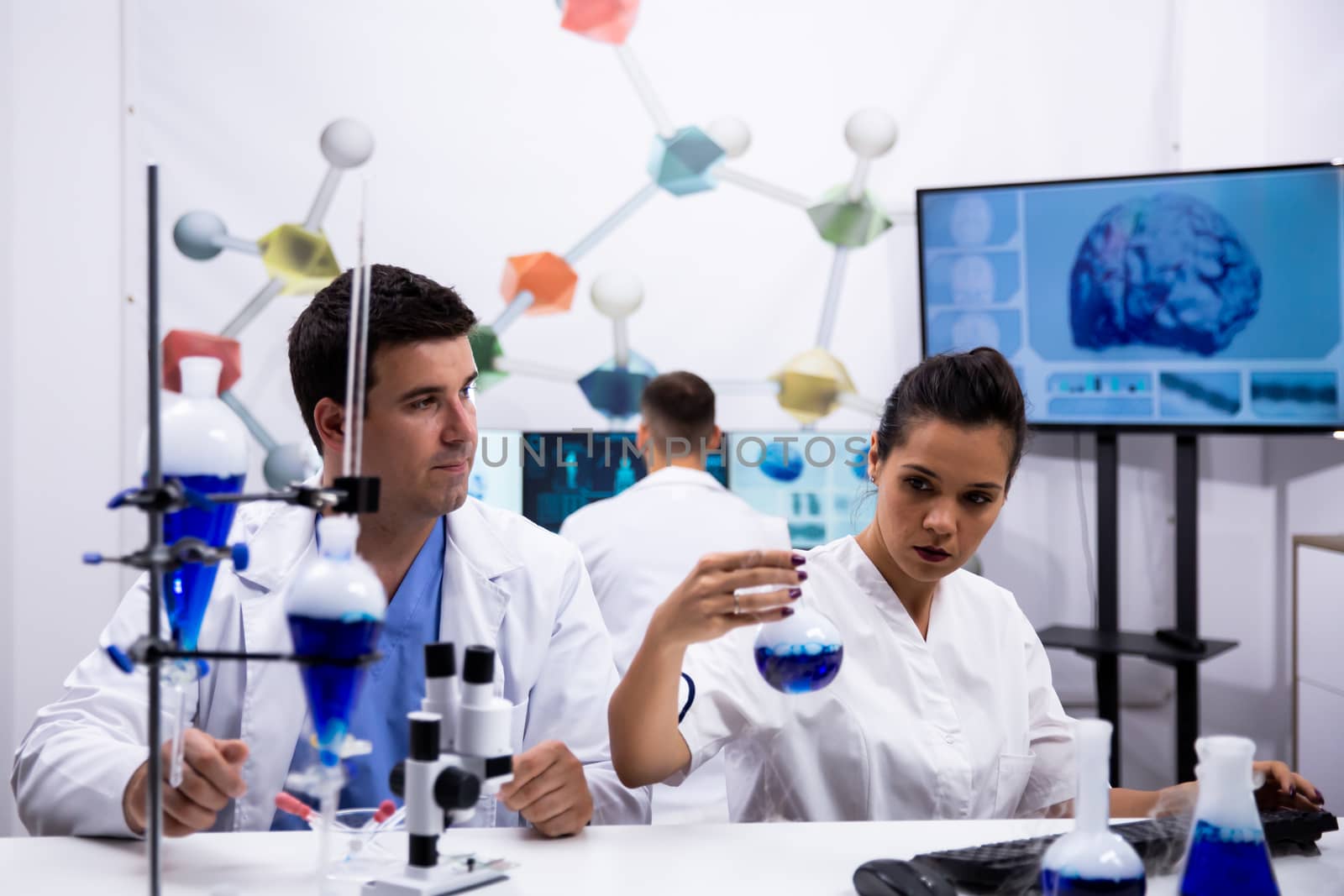 Young scientist wearing a white coat in research laboratory and assistant holding a test tube with blue liquid.