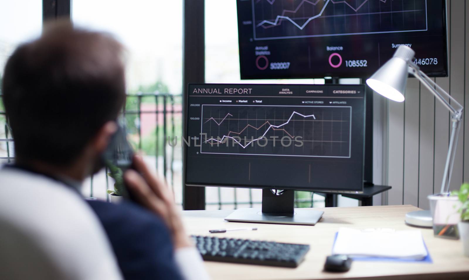Back view of young business man having a serious conversation on his mobile phone in his office. Graphics on monitor.