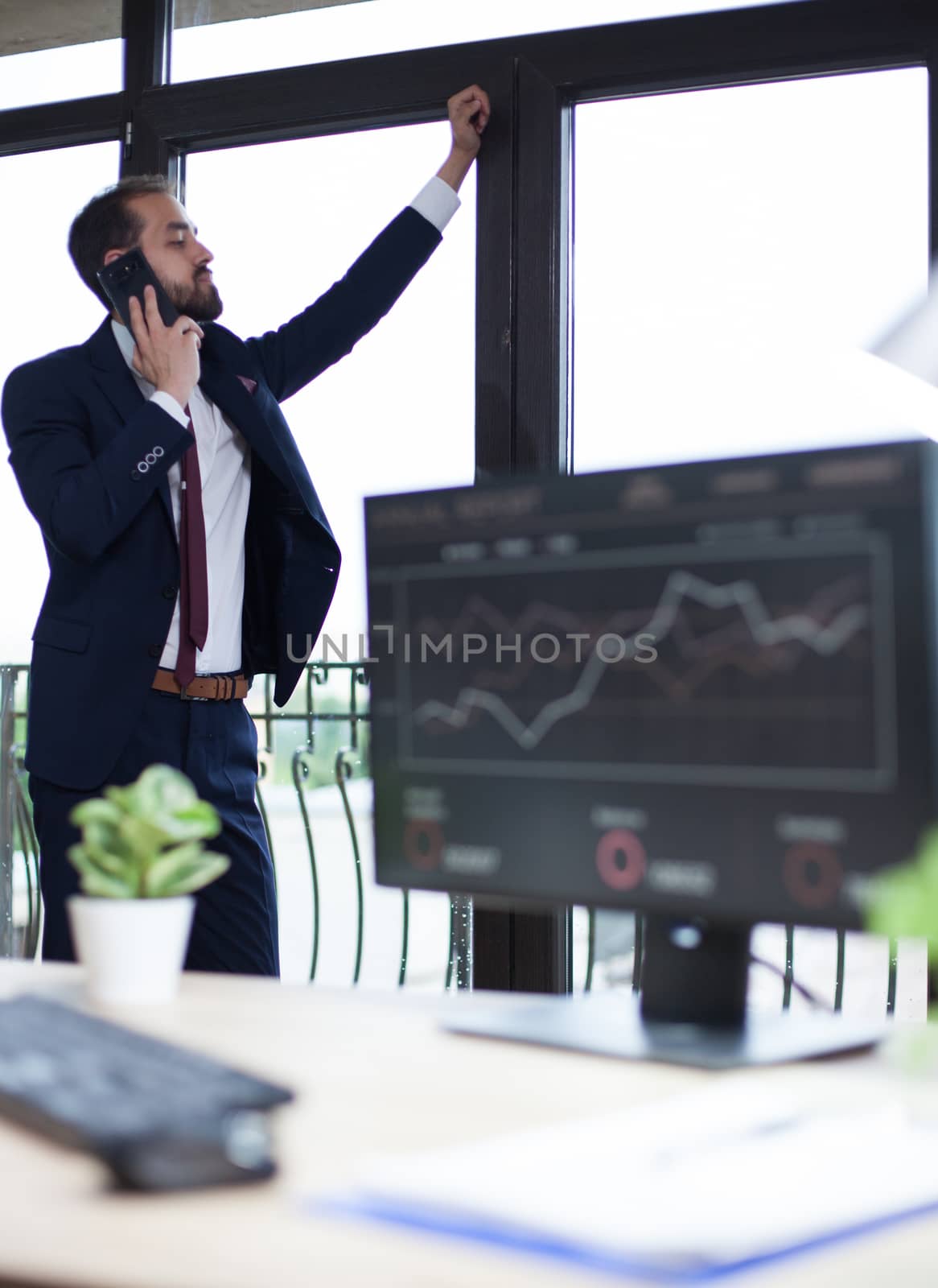 Confident young bearded businessman looking on the window and having a business conversation on the phone. Serious entrepreneur.