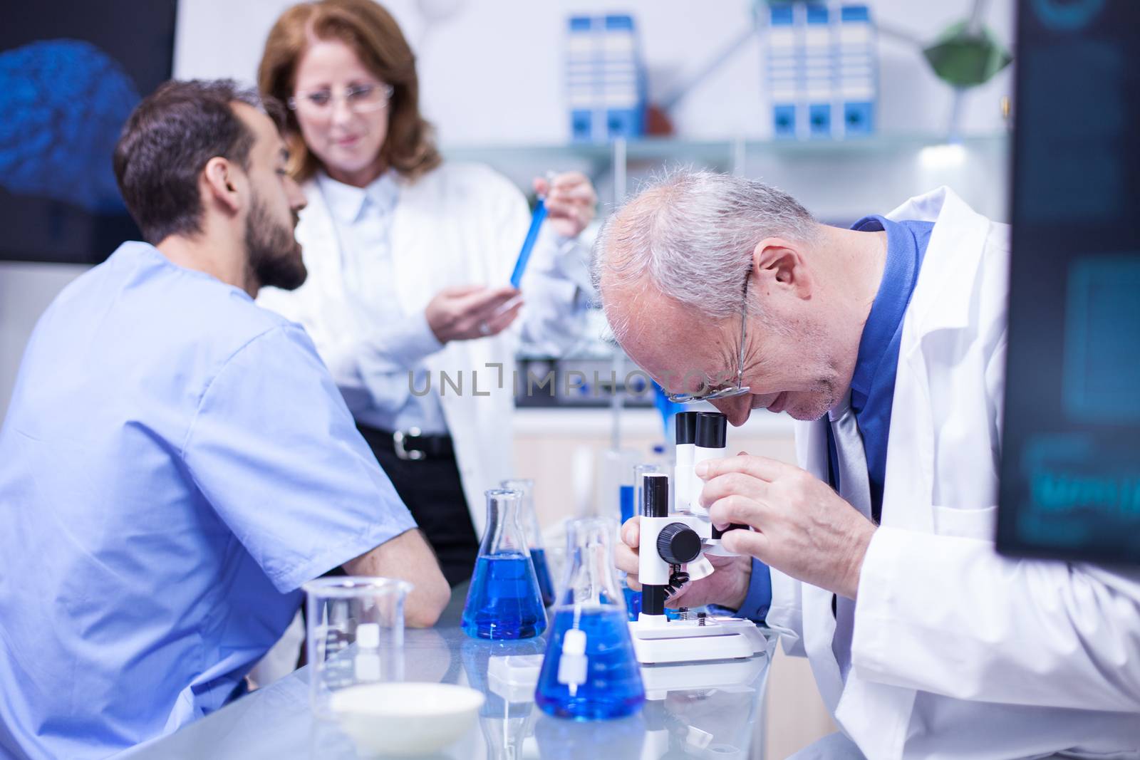 Senior scientist doing testing under the microscope in a modern laboratory. Team of scientist working in the background.