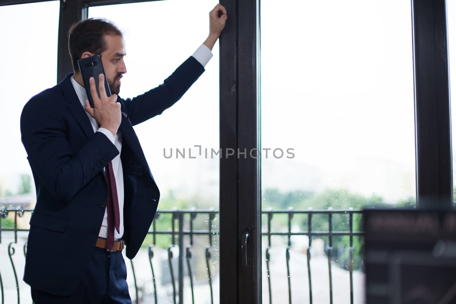 Handsome bearded businessman dressed elegant in his office looking on the window and talking on the phone. Serious entrepreneur.