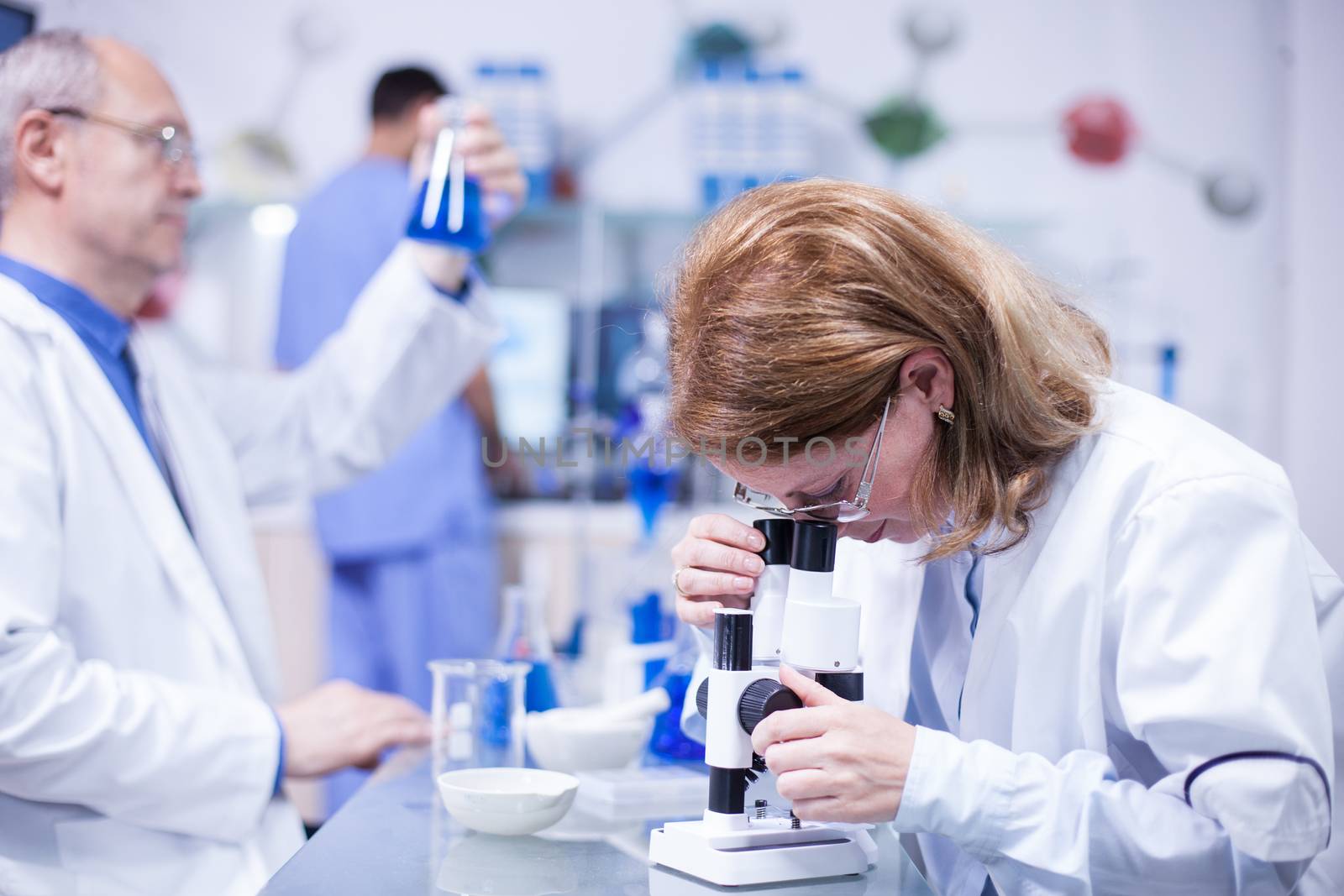 Middle age female chemist engineer working on a microscope in a laboratory. Senior scientist working with test tubes in the background.