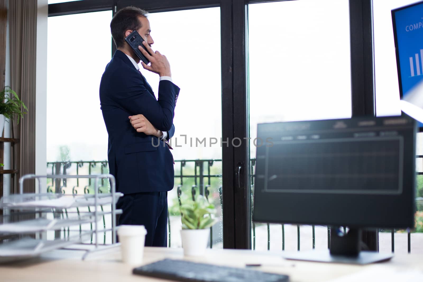 Young entrepreneur in suit having a serious conversation on his phone standing near the window. Computer monitor with graphics.