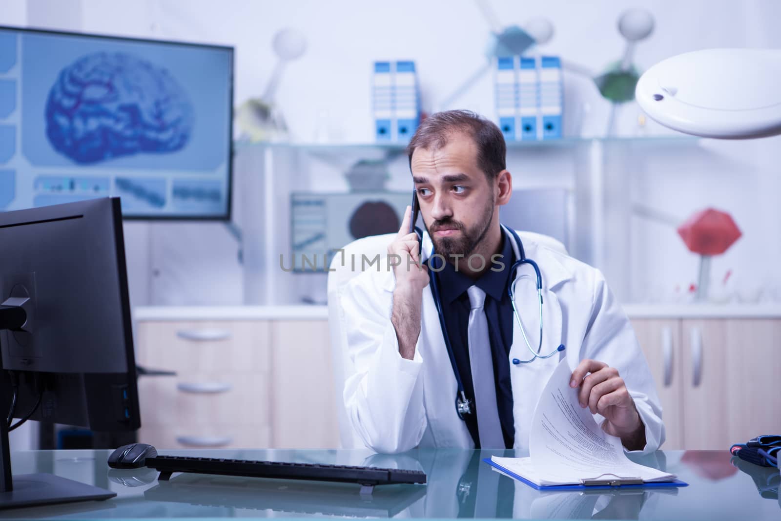 Male doctor using telephone while looking into his monitor from the hospital cabinet. Doctor checking the notes from his clipboard.