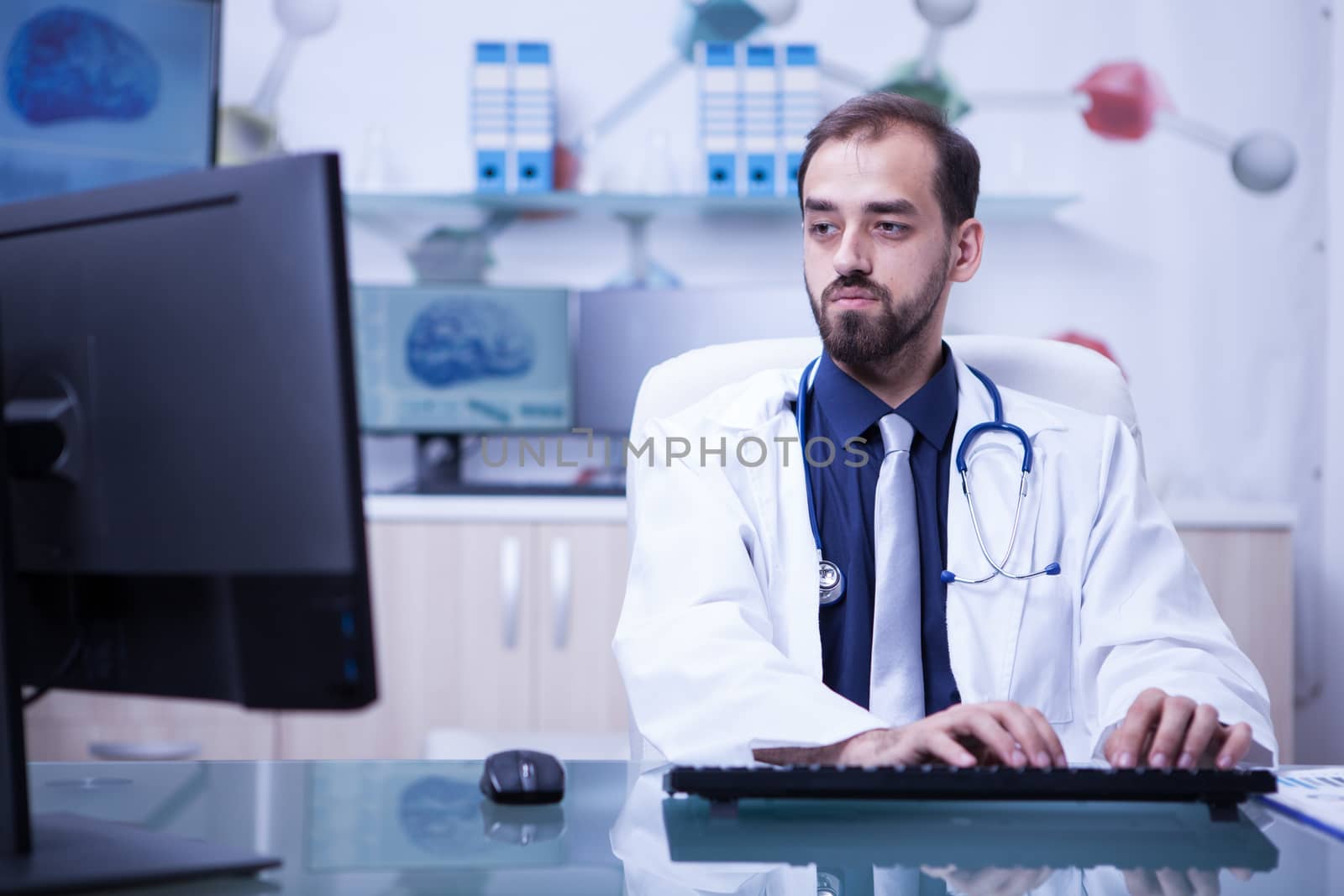 Young bearded doctor with both hands on the keyboard typing the recipe for a patient. Doctor concentrating on his work at the clinic.