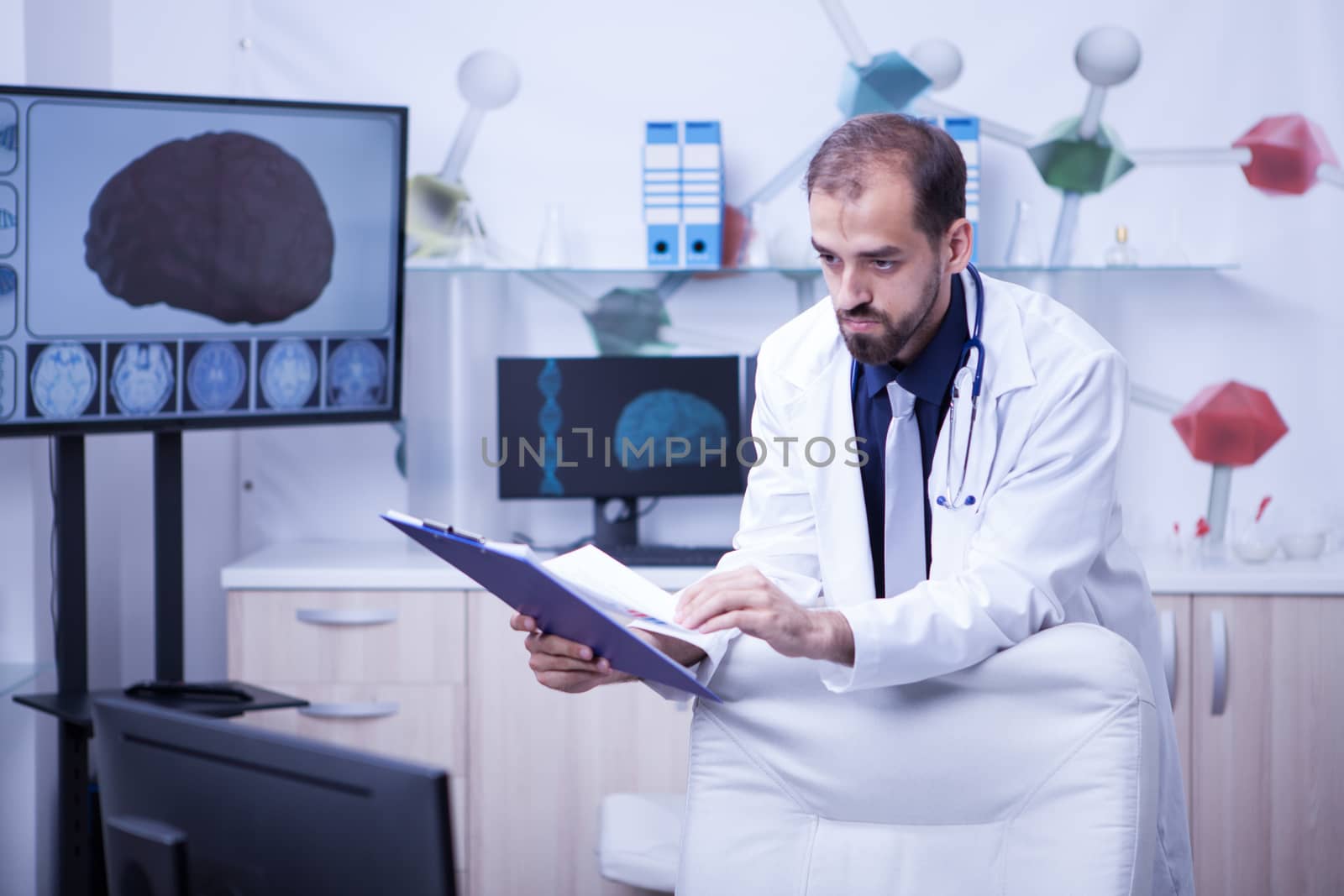 Young and handsome bearded man looking into his monitor and checking his hand notes from clipboard. Doctor in uniform.