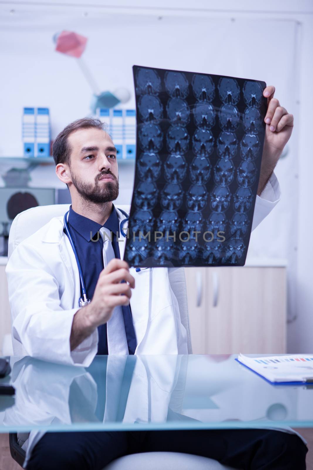 Young doctor at the clinic cabinet holding a brain radiography. Doctor checking the brain scan of a patient.