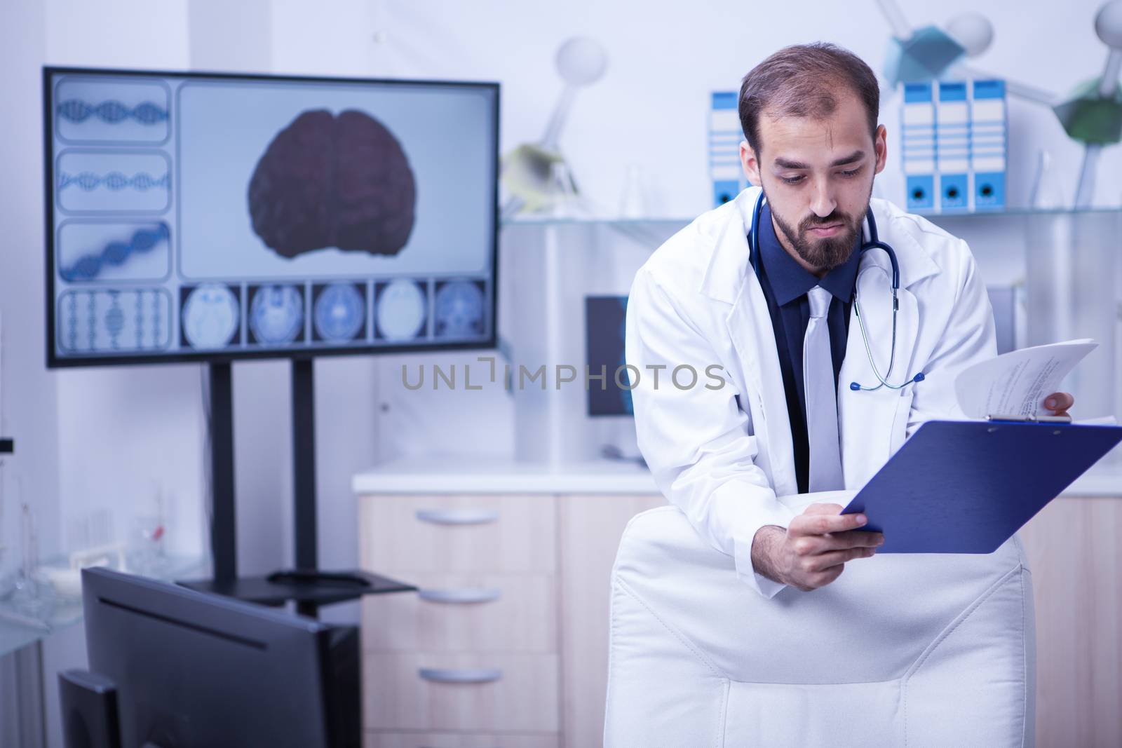 Man doctor in his cabinet from the hospital looking on his notes from clipboard. Brain on display on the background.