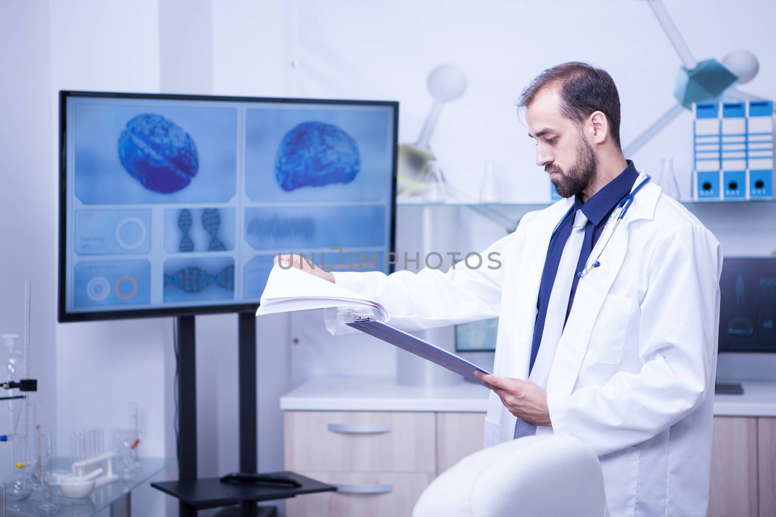 Young doctor turning pages with analyze on his clipboard in a laboratory for brain problems. Brain displayed on monitor.