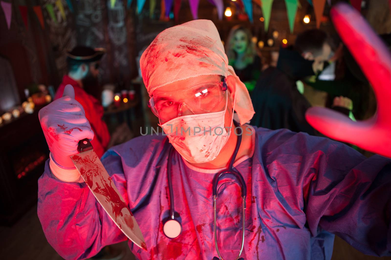 Creepy doctor with his covered holing a kinfe with blood and looking into the camera at halloween party. Halloween event.