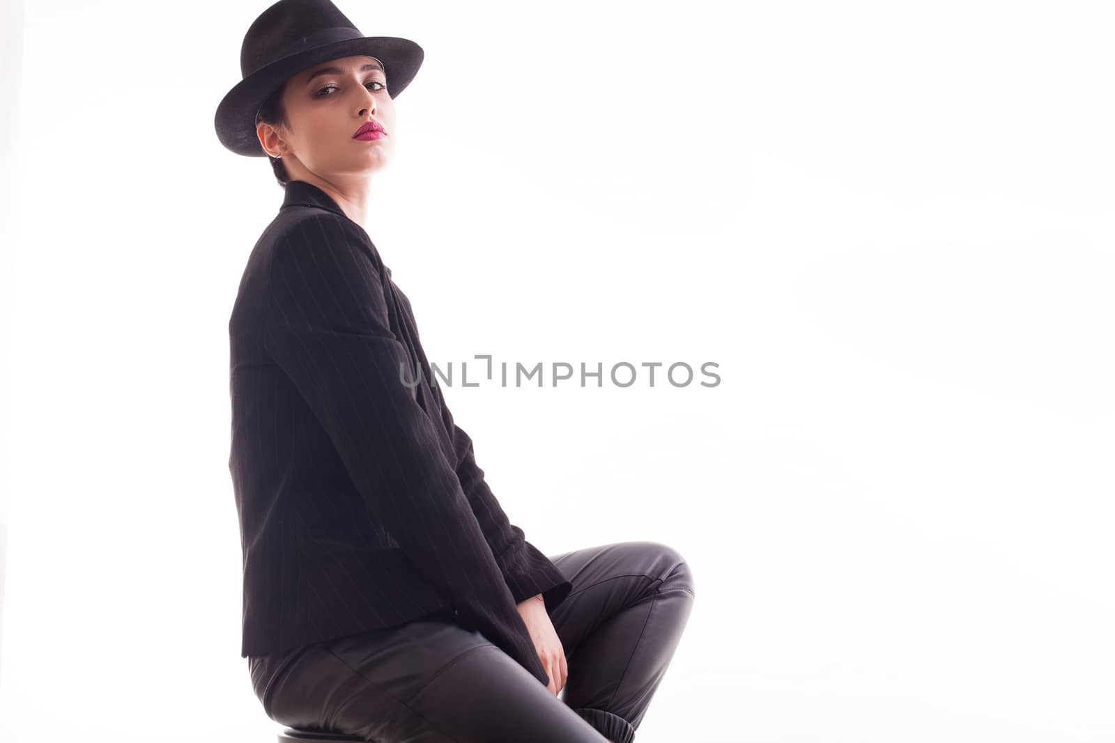 Young fashion model with a stylish hat and jacket posing sitting on a char in studio over white background. Beautiful young female in studio.