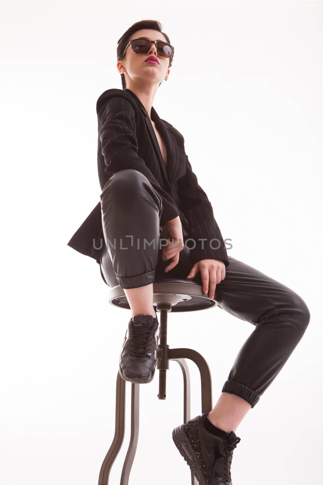Portrait of fashion model looking down on the camera sitting on a chiar in studio over white background. Full body portrait of stylish woman.