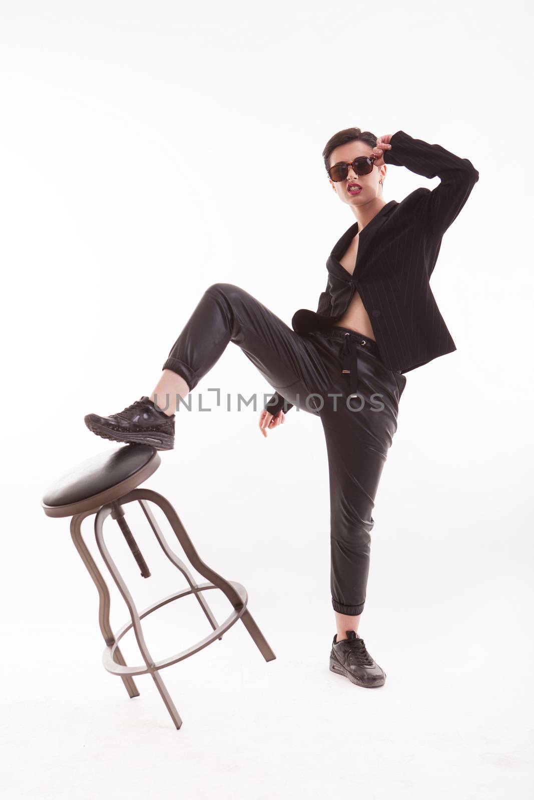 Stylish young fashion model posing with one leg on a chiar over white background in studio. Comfortable outfit.