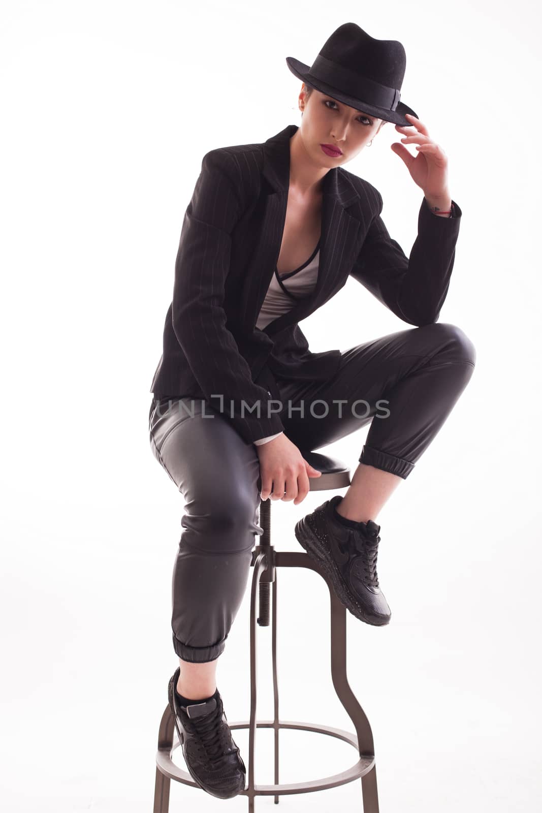 Beautiful caucasian young female sitting on chair and holding her stylish hat over white background looking at the camera. Elegance young woman.