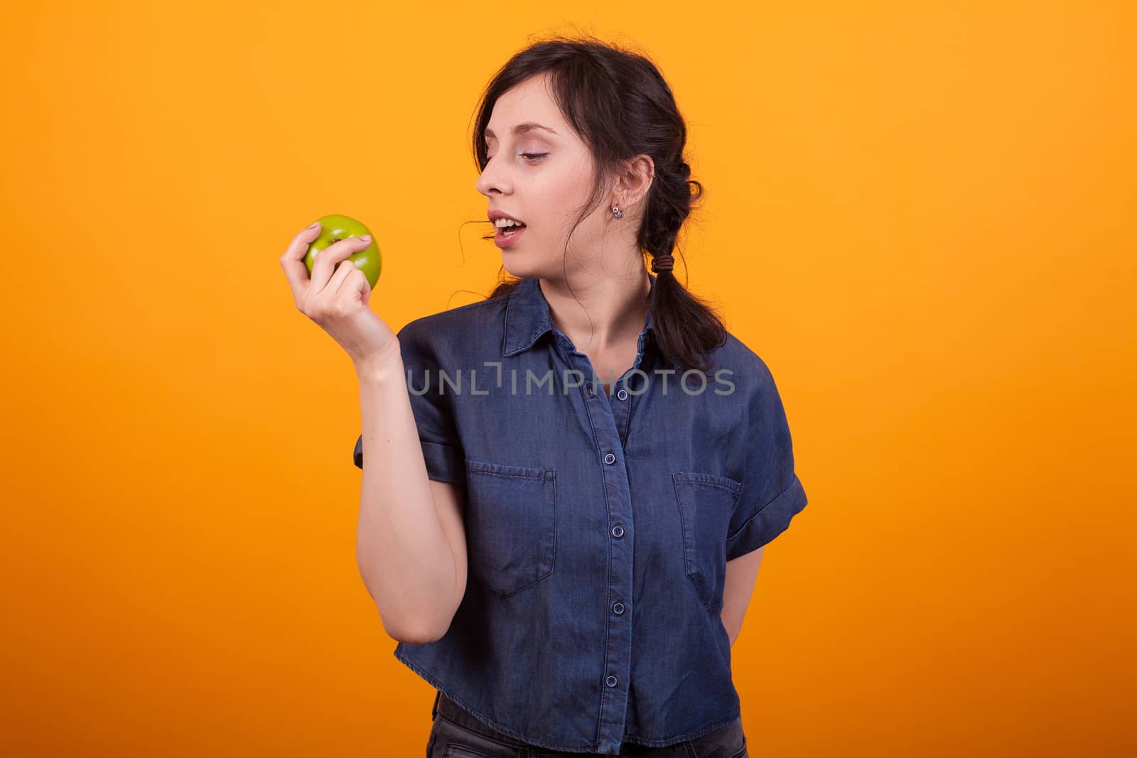 Side portrait of cheerful young woman holding and looking at a green apple in studio over yellow background. Woman promoting healthy lifestyle. Eating apples for skin care.