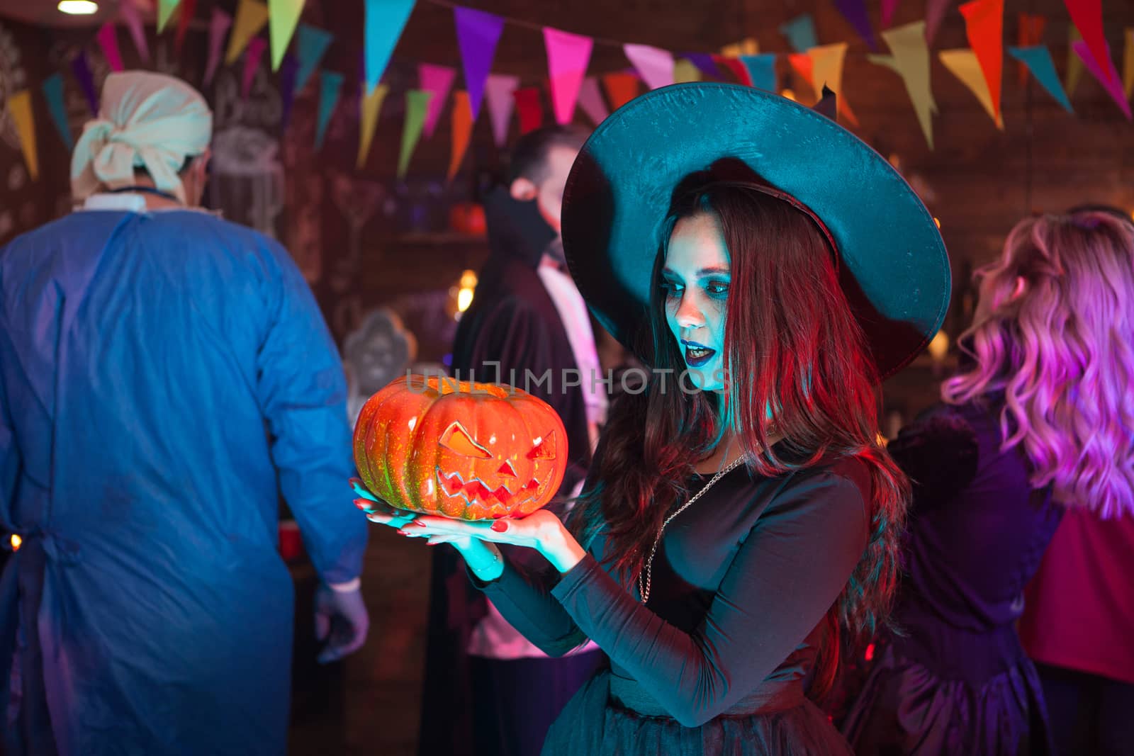 Portrait of young witch looking shocked at her punmpkin for halloween celebration. Woman with a big witch hat.