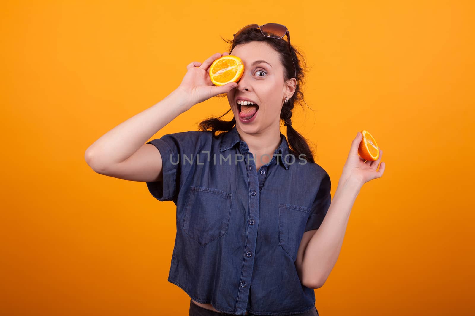 Portrait of a lovely young smiling and covering one of her eye with a fresh and tasty orange in studio over yellow background. Cute lady with delicious citrus. Beautiful woman holding sliced summer fruit.