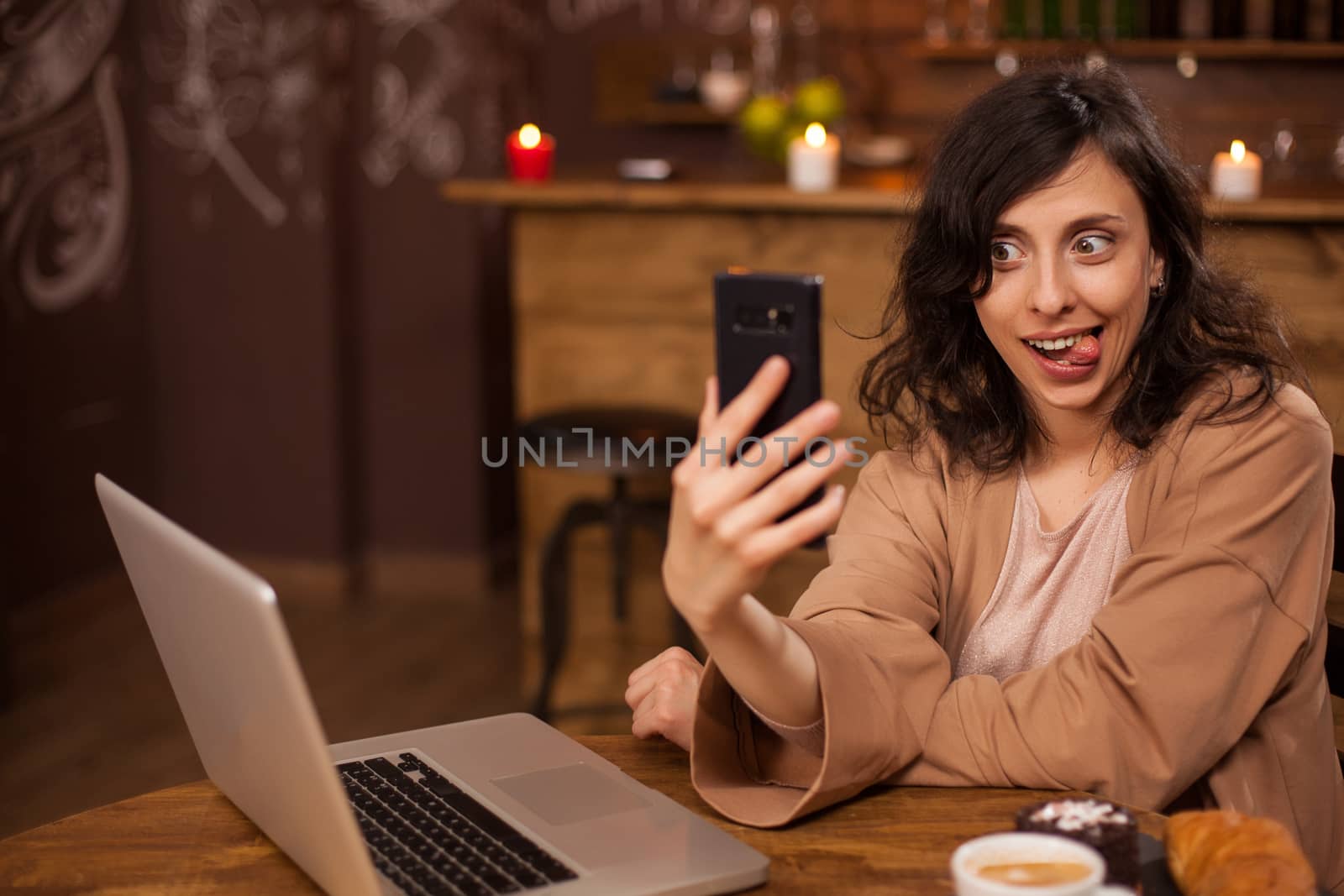 Smiling beautiful young woman taking funny selfie on her phone sticking her tongue out in a coffee shop. Girl with lapto near her. Funny lifestyle.