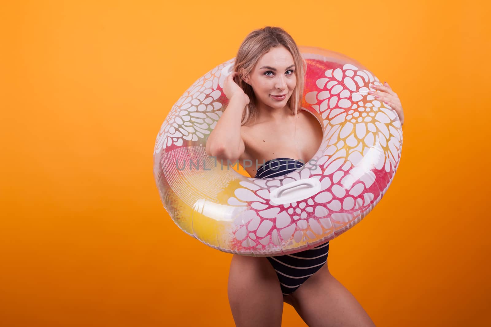 Thrilled beautiful woman with blue swimming suit playing with her hair and looking at the camera over yellow background. Sexy young woman in blue swiming suit inside a colorful swimming ring.