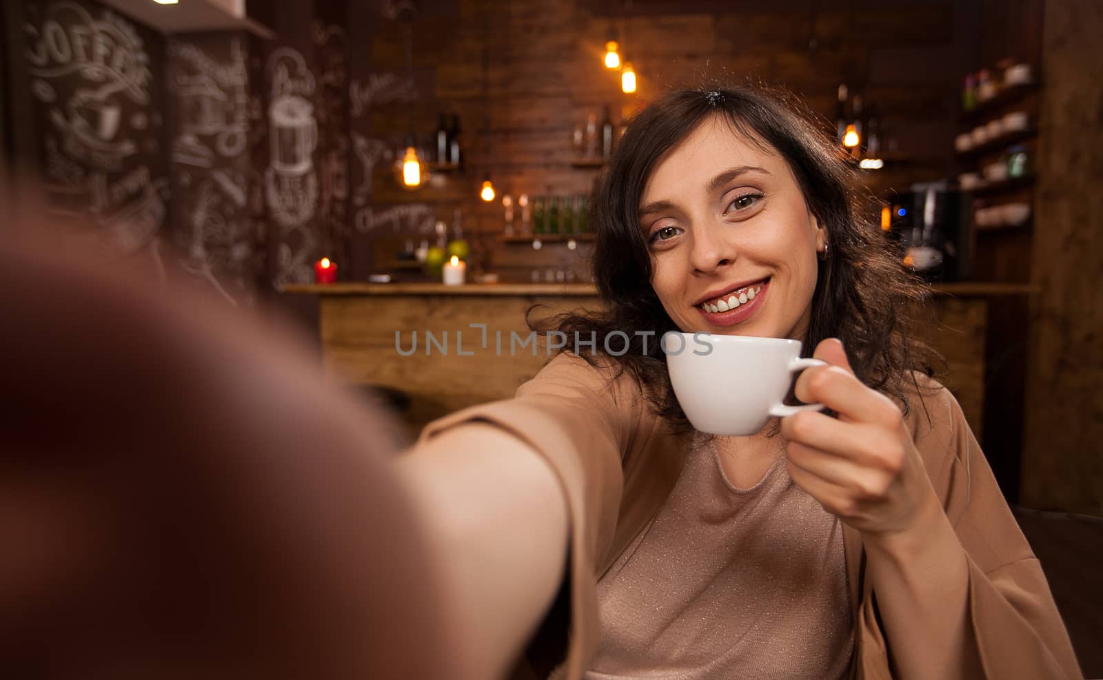 Beautiful woman using mobile phone to take a selfie during her work in a coffee shop. Pretty girl holding a cup of coffee. Young woman relaxing in a coffee shop. Casual day for a young woman.