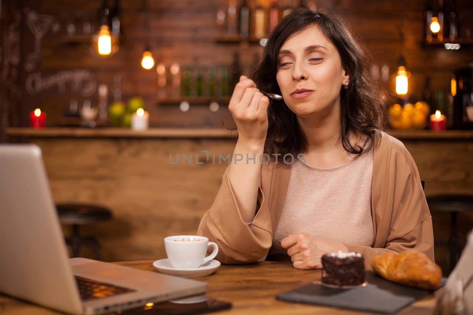 Portrait of beautiful woman enjoying a tasty cake in a coffee shopt with laptop near her. Attractive woman eating delicious dessert in coffee shop.