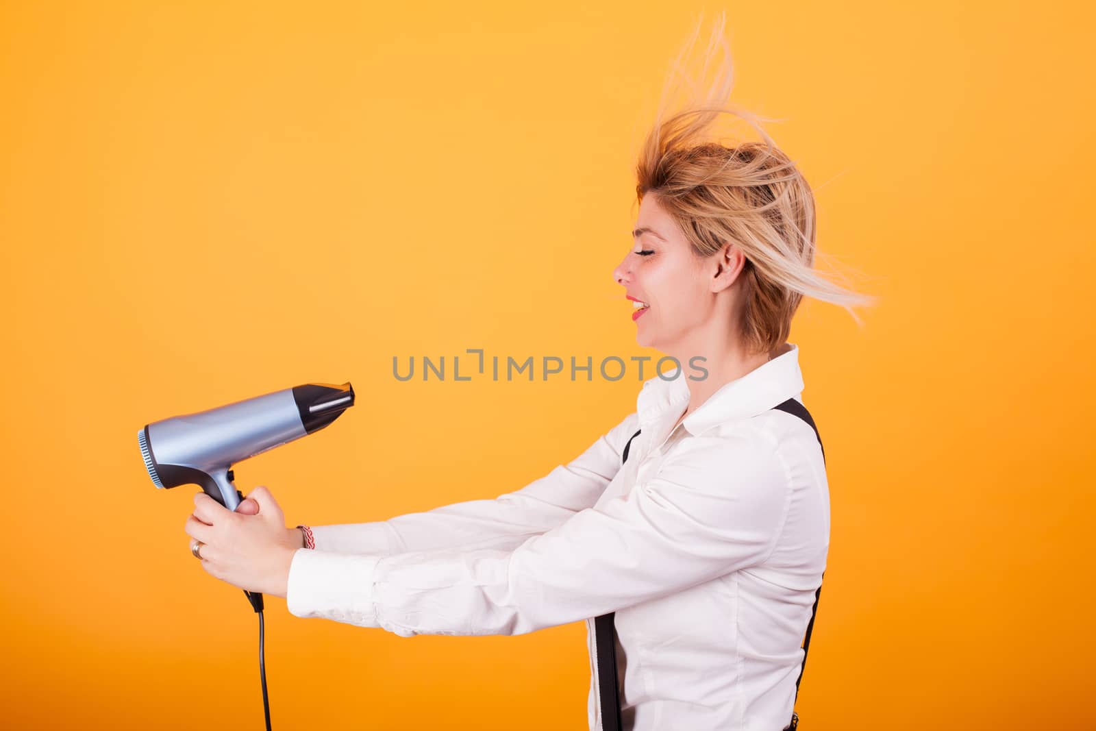 Young funny woman blowing her short hair with a hair drayer over yellow background. Cool hairstyle. Haircare.