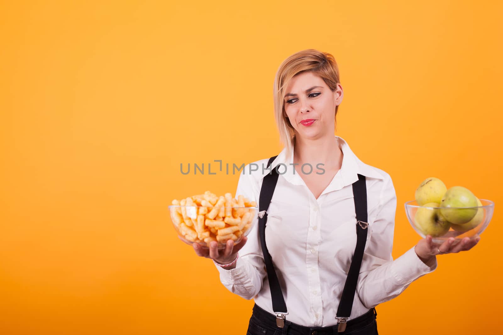 Pretty young woman holding two bowls of corn puffs and green apple over yellow background. Beautiful smile. Healthy food.