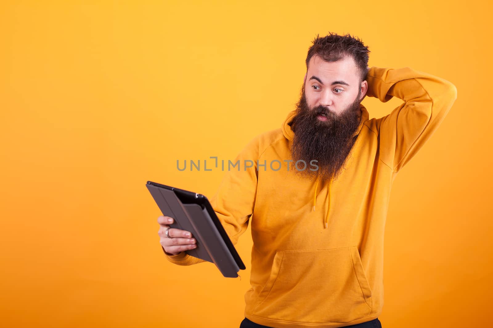 Handsome young man with long beard looking confused at tablet over yellow background. Handsome man.