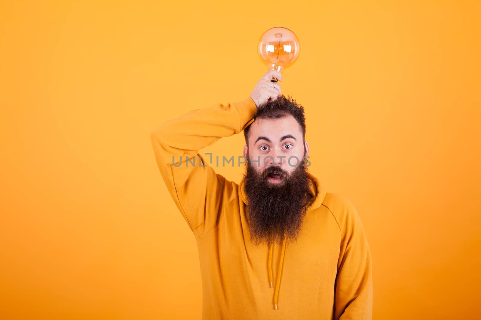 Hipster man with long beartd looking at the camera while holding a light bulb over his head. Smart solution. Handsome man.